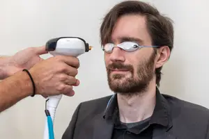 A man wearing glasses is being treated with a laser by another person in a room