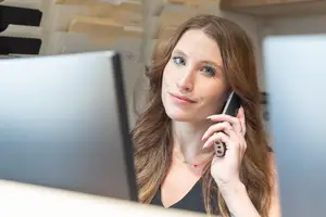 A woman with long hair wearing a black top and a necklace is sitting in front of a computer and talking on the phone