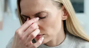 A woman with blonde hair is looking at something while her left hand is on her forehead. She seems to be in a room with a blurry background. She is wearing a gray sweater and has a small earring on her left ear.