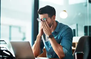 Man sitting at a desk with a laptop, looking stressed and covering his face with his hands
