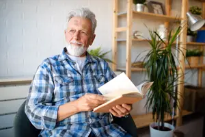 A senior man wearing a blue and white checkered shirt is sitting in a chair and reading a book.