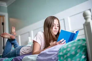A young girl is lying on a bed while reading a book and looking at something with her right hand