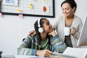 A young girl wearing headphones and a green shirt sitting in a chair with a woman standing behind her holding a coffee cup and smiling at the girl.