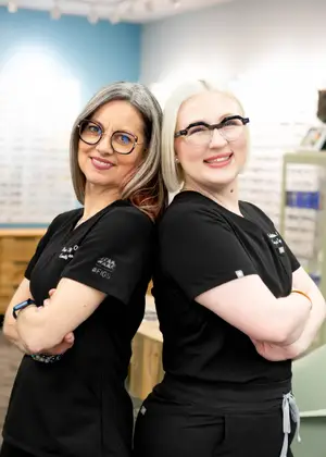 Two women in black glasses and shirts are smiling and posing for a photo in a room with blue walls, a desk, and a cabinet.