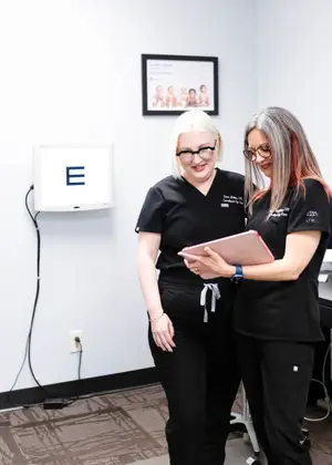 Two women in black scrubs, one wearing glasses and the other wearing a watch, standing in a room with a white wall, a picture frame, and an electronic device on the wall, they are looking at a tablet. 