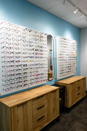 A room with wooden cabinets, a mirror, and a wall displaying various eyeglasses. The room has a carpeted floor, a blue wall, and a white ceiling with ceiling lights. The eyeglasses are displayed on a white wall with a grid pattern. The cabinets have drawers and handles, and the mirror is attached to the wall.