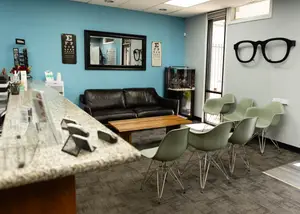 An empty optometrist office with blue walls, a black leather couch, and green chairs, with a fish tank and a glass counter with a monitor and glasses on it.