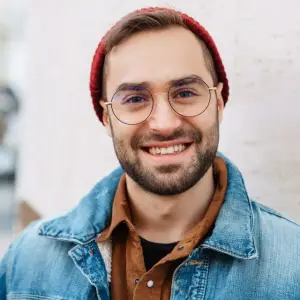 A smiling man with a beard and glasses wearing a denim jacket and a red beanie