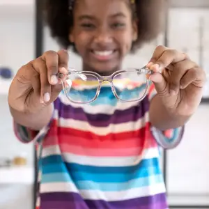 A girl in a colorful striped shirt is smiling while holding a pair of clear glasses.