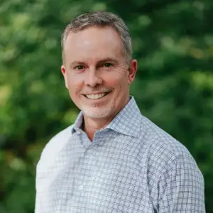 A man with a beard and a mustache is smiling in front of a blurry green background with trees.