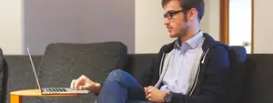 A man sitting on a couch wearing glasses and a jacket working on his laptop