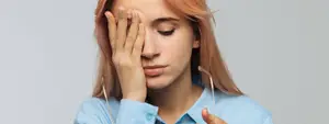A woman with red hair is touching her forehead with her left hand while holding a pair of glasses in her right hand. She is wearing a blue long-sleeve shirt and is probably in a room with a white wall.