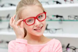 A young girl wearing red glasses smiles while posing for a photo in an optical shop