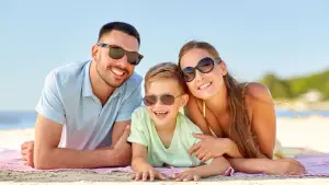 A man, a woman, and a boy are lying on a beach with sunglasses on, smiling and posing for a photo