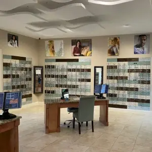 Inside an optical store with a desk and monitors, a person sitting on a chair, and glass walls with posters of people.