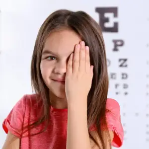 A young girl in a red shirt is smiling and covering her left eye with her hand, likely at an eye doctor's office.