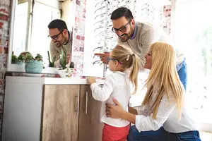 A family of three people is looking at eyeglasses on a mirror in a room with a brick wall and a white cabinet.