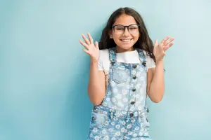 A girl with glasses and a white shirt poses against a blue background with her hands raised and smiling.