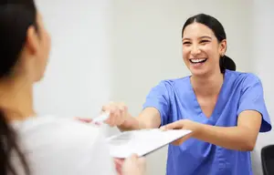 A smiling technician in a blue uniform is handing a paper to a patient in a white shirt in an indoor room.