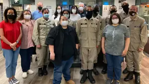 A group of people, including two women in pink and red tops, are standing together in a room. They are all wearing masks and appear to be in uniform.