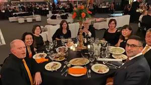 A group of people are seated around a table in a formal setting with plates of food, wine glasses, and a centerpiece of flowers.