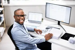 A smiling man sitting at a desk with a laptop and calculator in an office.