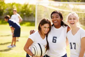 Three girls on a soccer field smiling at the camera, with a soccer ball in hand and a soccer goal in the background