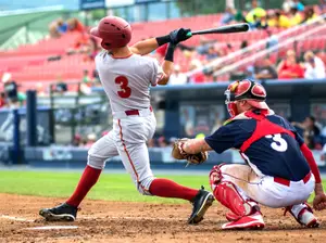 A baseball player wearing a number 3 jersey swings his bat while the catcher crouches behind him, both wearing helmets and gloves, on a baseball field with spectators in the background.