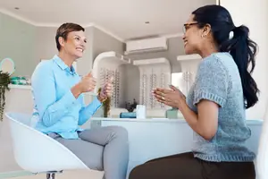 A woman wearing glasses is talking to another woman with a ponytail in a room with an air conditioner, plants, and a table with various items