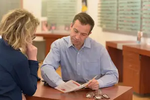 A man reads a brochure while sitting at a desk with a woman looking on, both in a professional setting.
