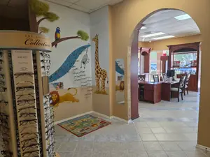 A room with a table, chairs, computer, shelves, and a carpet on the floor, with a wall featuring a giraffe, lion, and other animals, and a display of eyeglasses labeled "The Exclusive Collection".