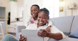 A smiling woman is sitting on a couch with a child on her lap, looking at a tablet, with a kitchen in the background.