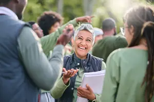 A woman holding papers is smiling and talking to a group of people in a forest.