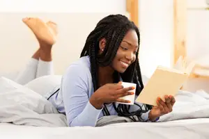 A woman is lying on a bed with her feet up, smiling and holding a book and a cup.