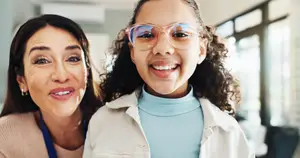An adult woman and a young girl with glasses smile at each other in a hallway