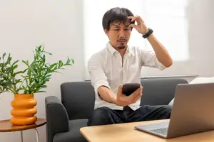 A man in a white shirt is sitting on a couch looking at his phone with a confused expression.