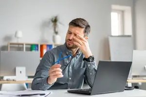 A man is sitting at a desk with a laptop and papers, holding a pair of glasses and touching his forehead with his right hand. He is probably feeling tired and stressed.