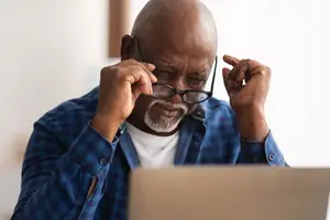 An elderly man wearing glasses and a blue shirt is sitting in front of a laptop, possibly working or browsing the internet.