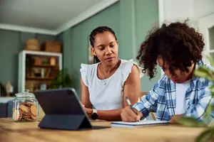 A woman and a child are sitting at a wooden table with a laptop and a notebook. The woman is wearing a necklace and a watch, and the child is holding a pen. The table has a jar of cookies, a glass jar, and a shelf with storage boxes. The wall is painted green, and there is a white door behind them.