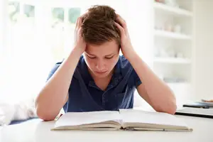 A young man with his hands on his head is sitting at a desk and reading a book.