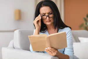 A woman wearing glasses is sitting on a couch reading a book and holding her head with her right hand