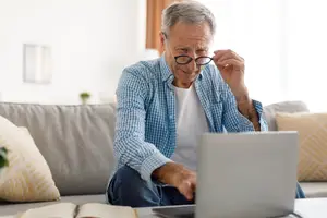 An elderly man sitting on a couch and using a laptop, wearing glasses and holding his chin with his right hand.