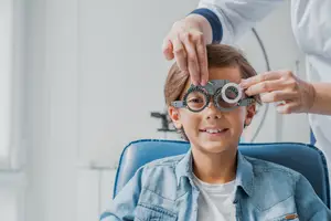 A young boy is sitting in a blue chair and wearing eyeglasses while a person in a white lab coat is adjusting his eyeglasses.