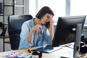 A woman sitting at a desk, rubbing her forehead and holding a pair of glasses, perhaps feeling stressed.