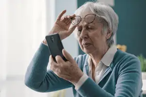 An elderly woman with white hair wearing glasses and holding a smartphone in her hands while looking at the screen