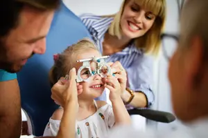 A young girl wearing glasses is being examined by an adult man and woman, with the woman holding the glasses and smiling.