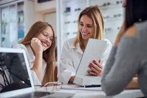 Two women sitting at a desk in a retail store wearing white lab coats looking at a piece of paper with a smiling girl standing next to them.
