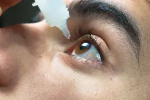 A close up of a woman applying eye drops to her eye.
