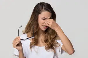 A woman is holding a pair of glasses and covering her nose with her right hand, probably sneezing.