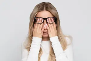 A woman with blonde hair covering her face with her hands and wearing glasses.